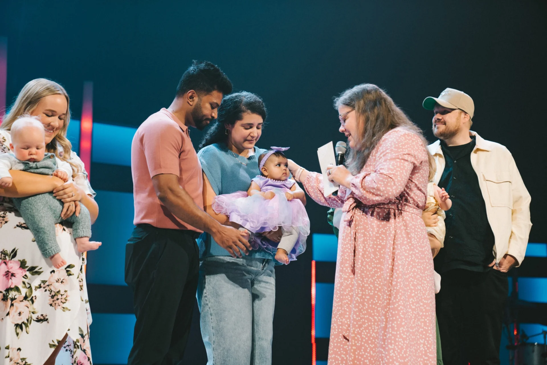 Families holding their babies on stage during a child dedication, as a pastor with a microphone blesses a baby in a purple dress.