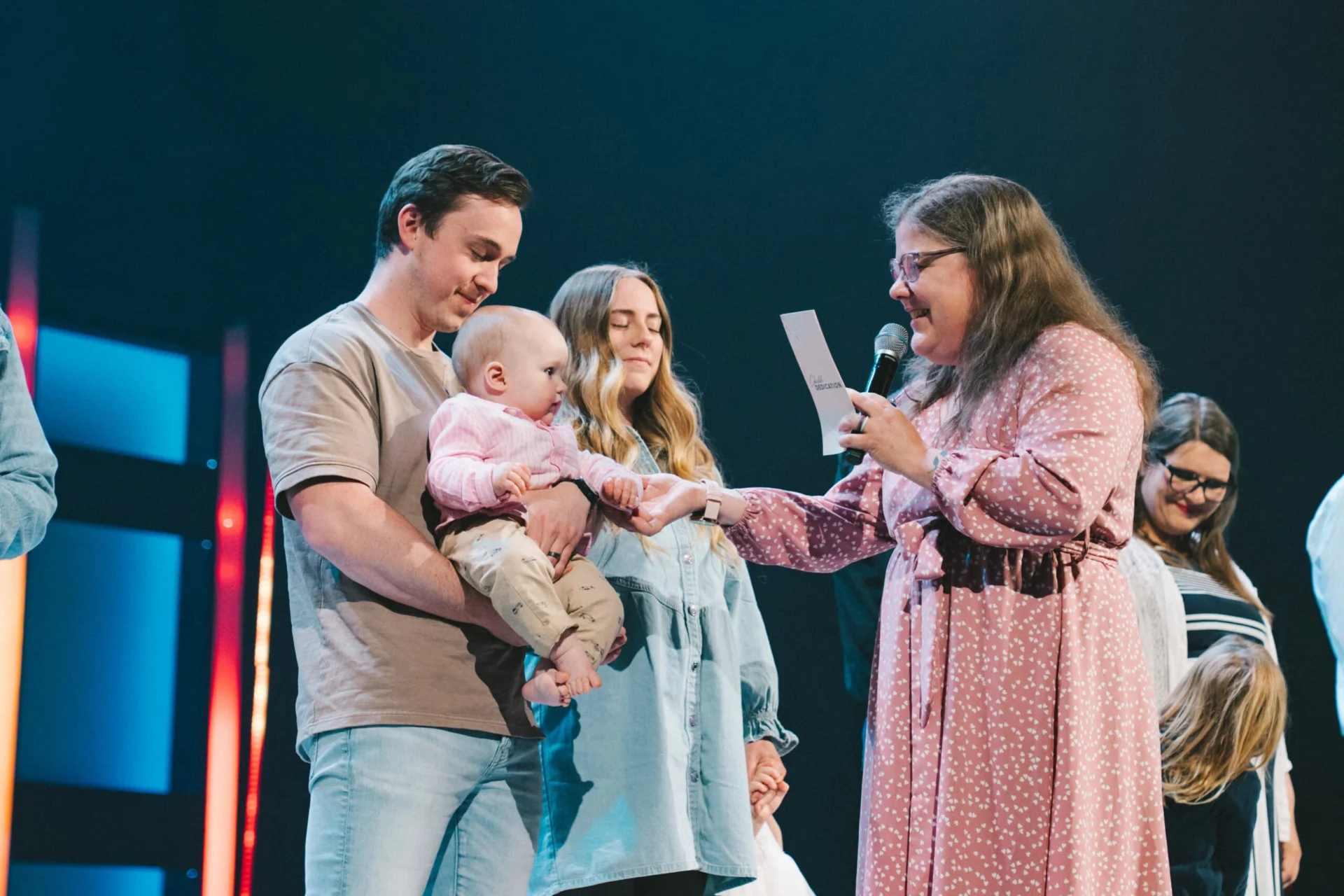 A father holds his baby on stage while a pastor reads the dedication and holds the baby's hand.