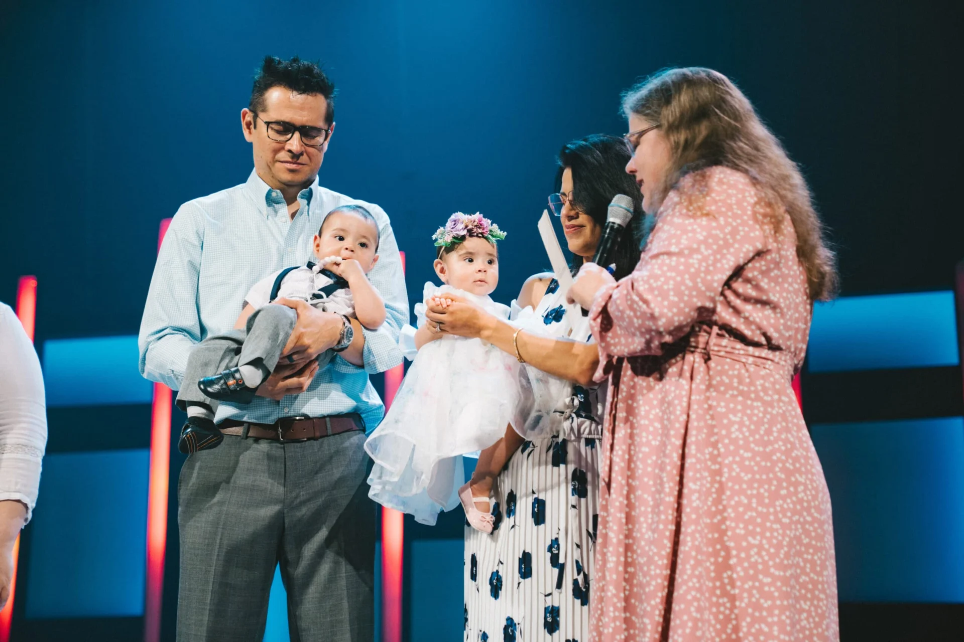 Two families hold their babies on stage, one in suspenders and one in a white dress with a flower headband, during a dedication.