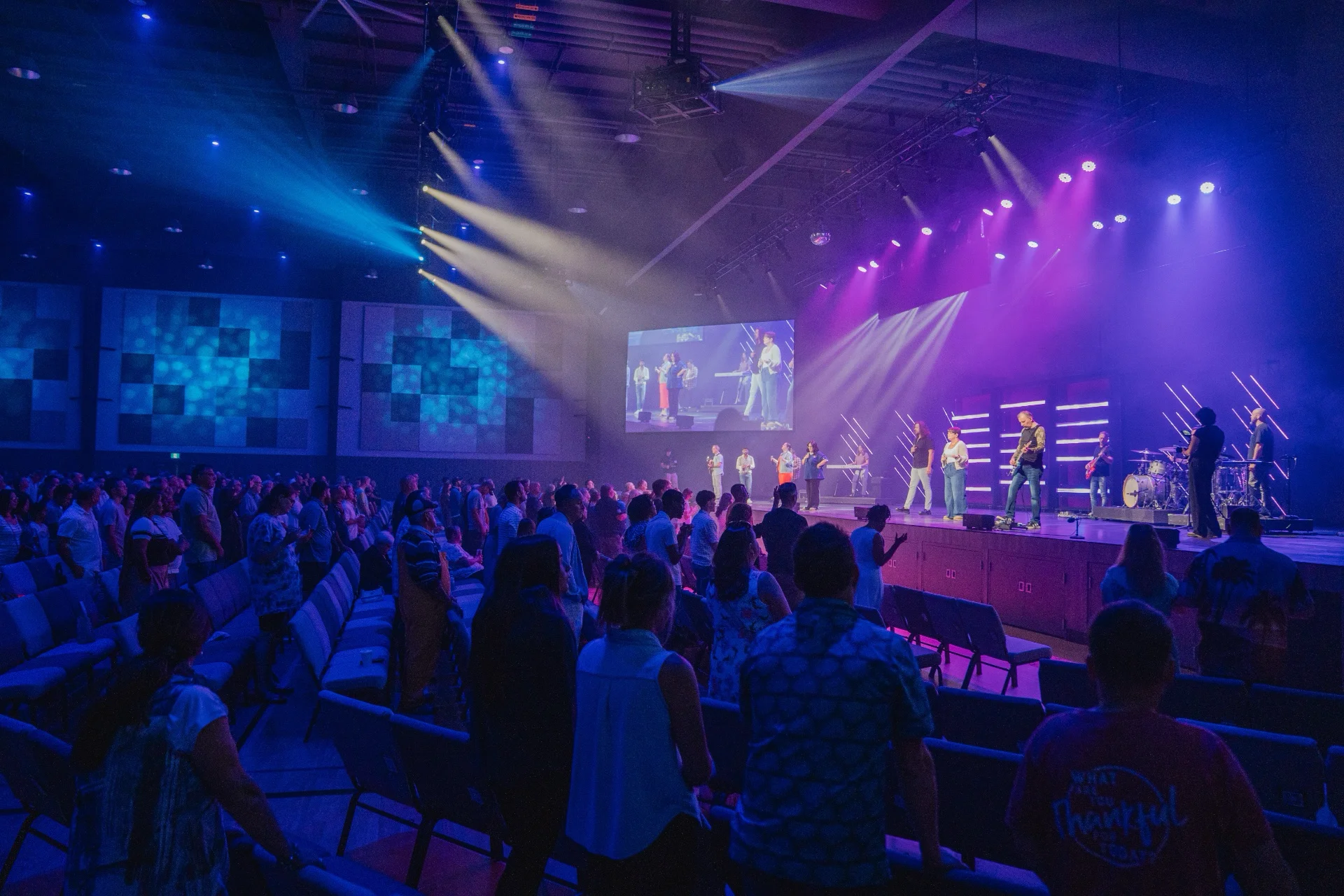 A full auditorium of people standing in worship during a Sunday Experience, with the band on a brightly lit stage.