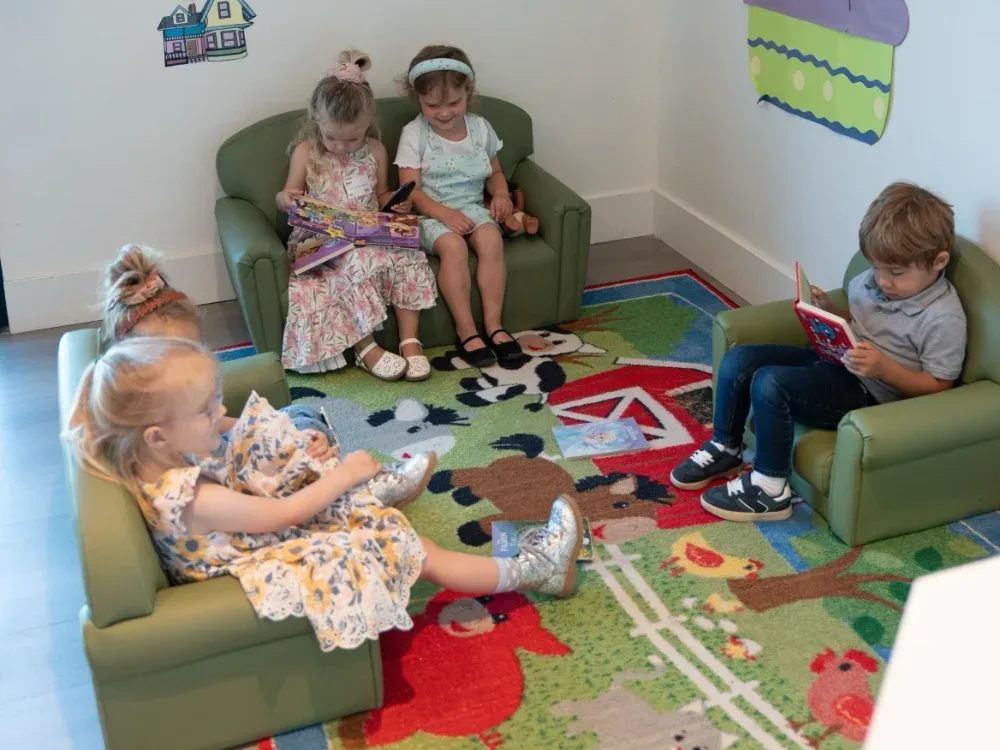 Young children reading picture books on small couches in a decorated kids room with a farm rug.