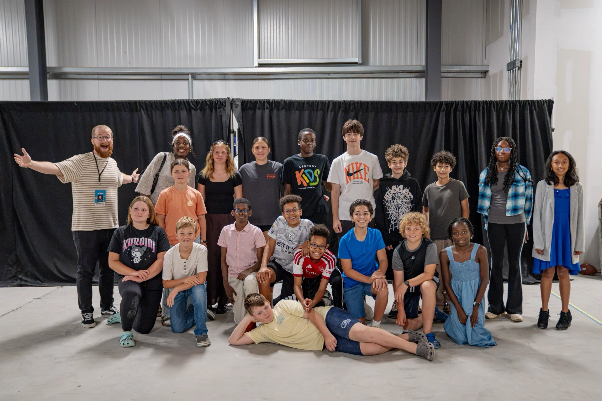 A large group of middle school students standing and kneeling together for a group photo in a big open room with a black curtain backdrop, with a leader at the side.