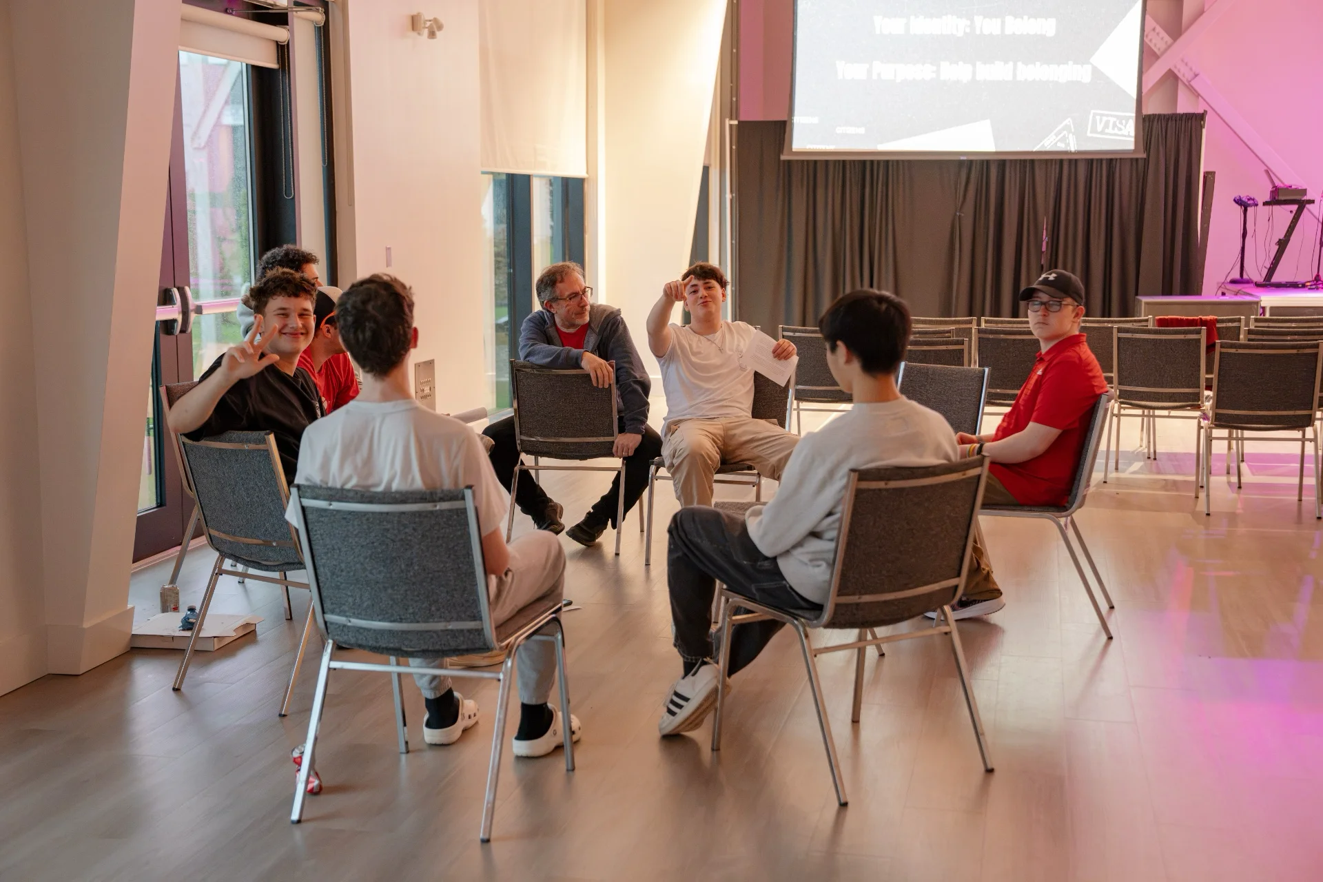High school students and an adult leader sitting in a circle of chairs in a bright room, one student holding a page and talking while the others listen.
