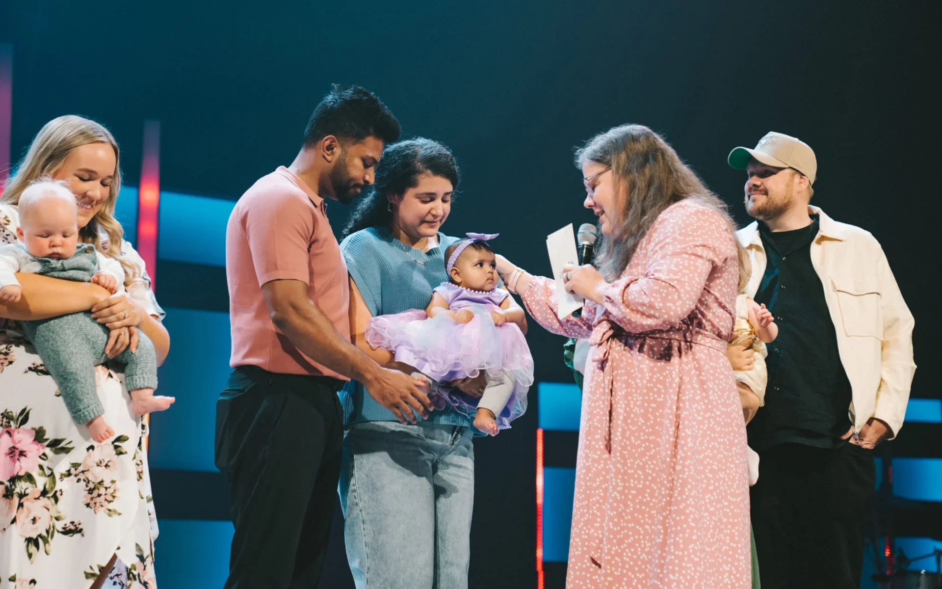 Parents holding their babies on stage during a child dedication, with a leader reaching toward one of the children.