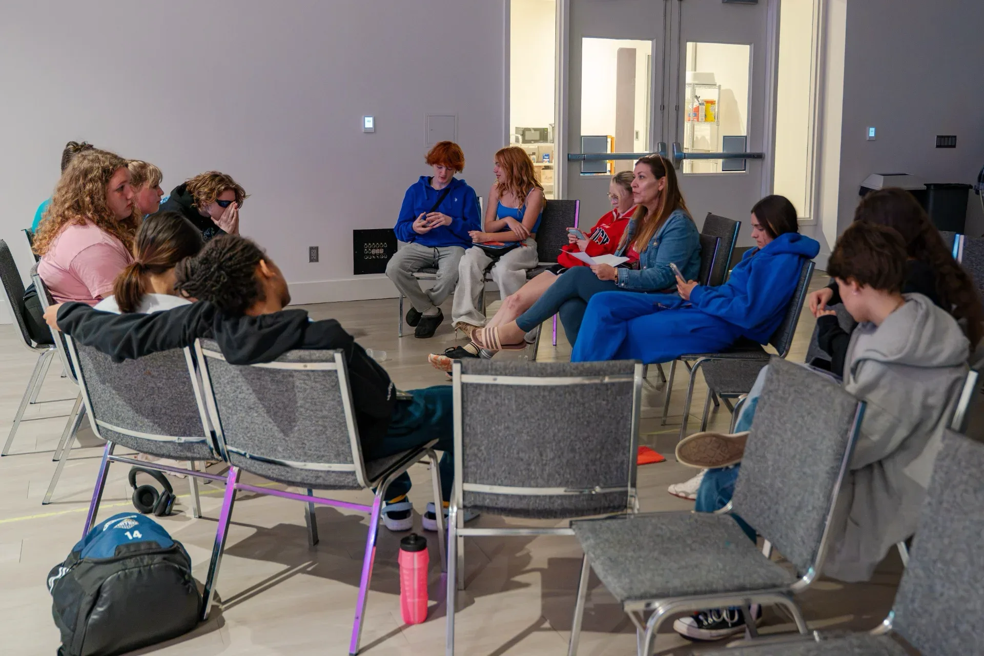 High school students and a leader sitting in a circle of chairs talking in a bright room.